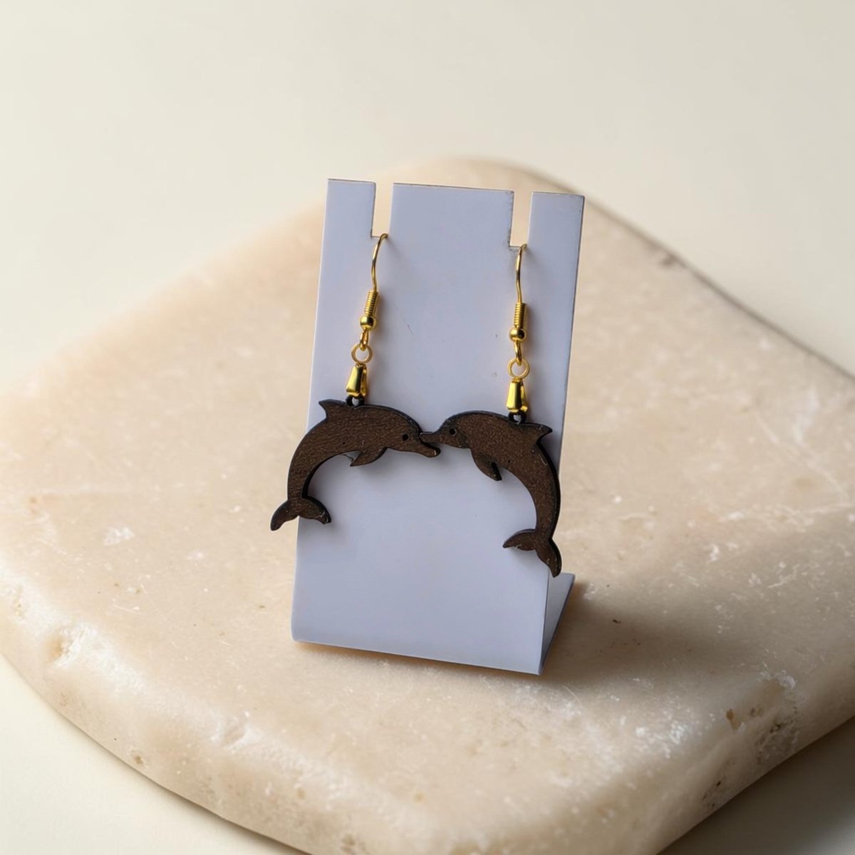 The Dolphin Earrings | Lasercut Walnut Wood by Mirabilia Boutique are displayed on a white stand atop a beige stone, with the earrings facing each other for a symmetrical, ocean-inspired charm.