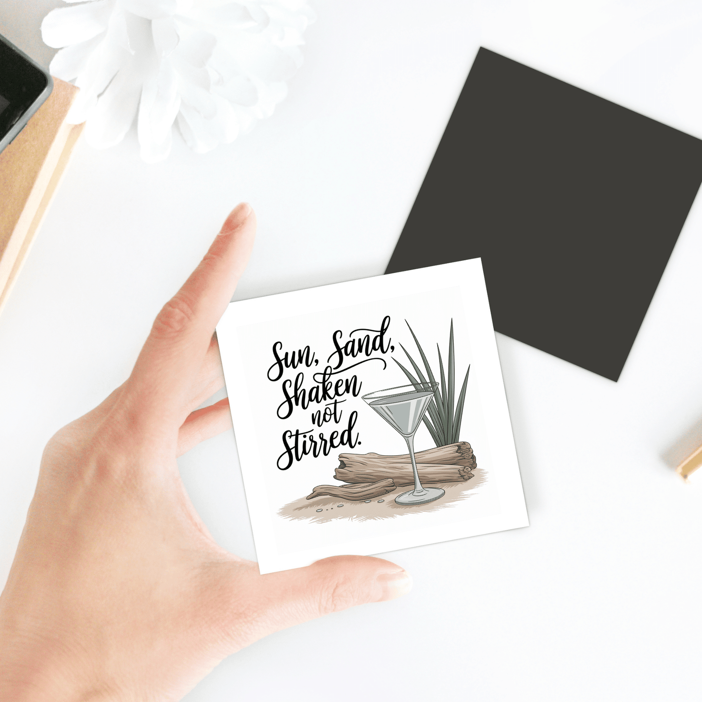 A hand holds the Sun, Sand, Shaken Not Stirred Magnet – Coastal Cocktail Quote by Mirabilia Boutique, featuring a martini glass with driftwood and plant—perfect for cocktail lovers or beach homes. "Sun, Sand, Shaken not Stirred" is printed on the card.