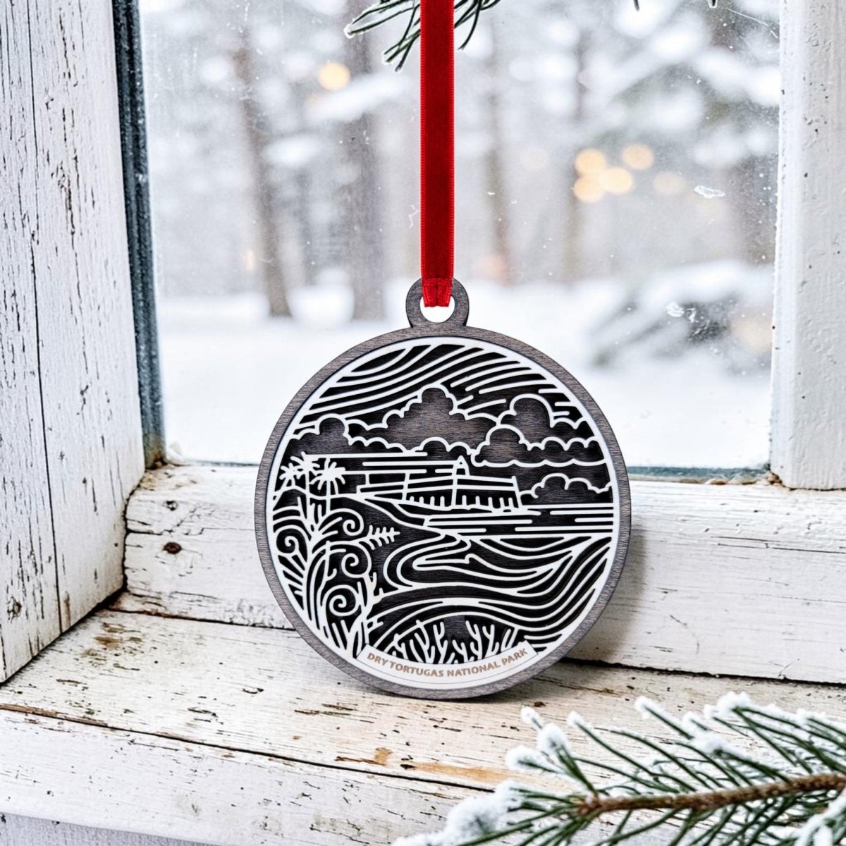 A Mirabilia Boutique Dry Tortugas National Park Christmas Ornament, featuring a silver landscape design with intricate lines illustrating nature and a distant building, hangs on a snowy window by a red ribbon. Frosted pine needles can be seen in the foreground.