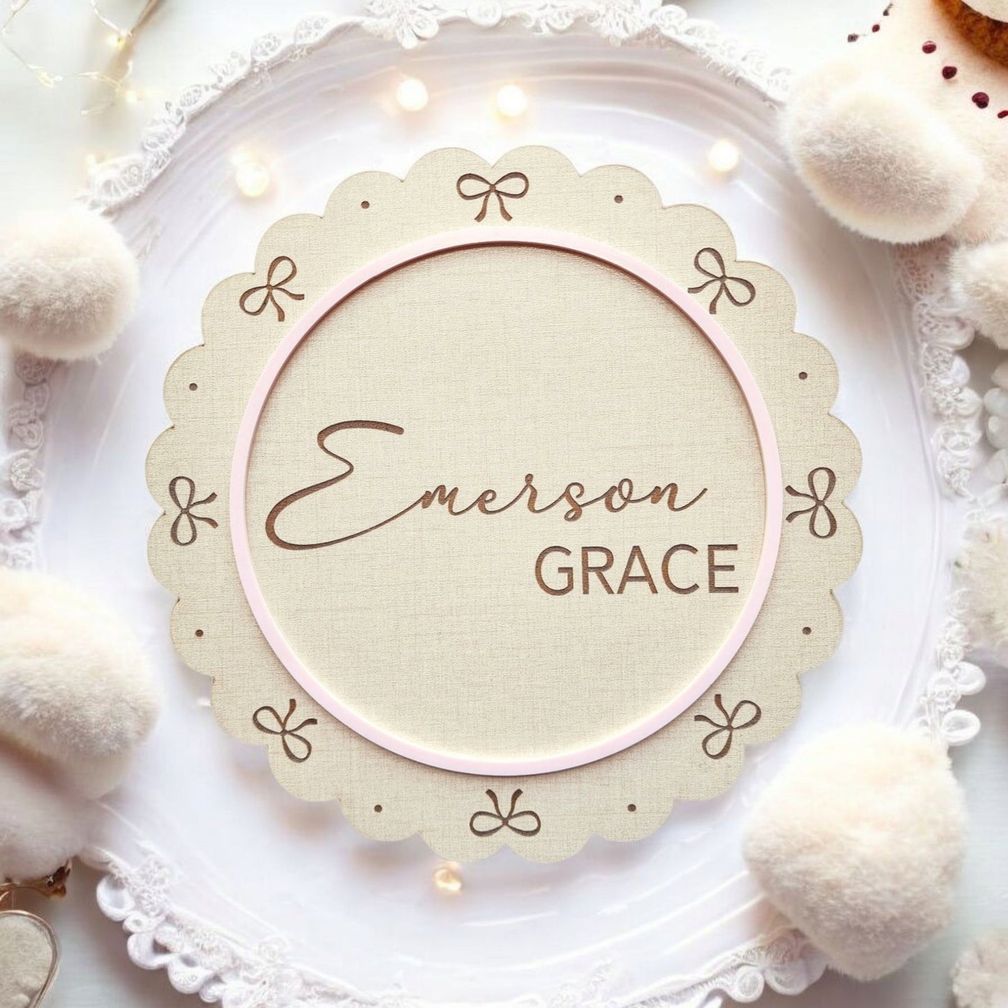 The Personalized Baby Name Shelf Sign by Mirabilia Boutique features scalloped edges, bows, and pink trim with "Emerson Grace" in cursive and bold. Ideal nursery shelf decor or a custom newborn gift, it’s accented with lace, cream pompoms, and fairy lights.