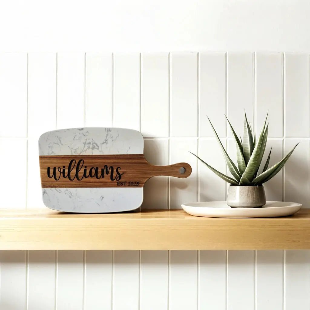 The Personalized Marble and Acacia Serving Board by Mirabilia Boutique, custom-engraved with a name, leans beside a potted succulent on a wooden shelf, creating a stylish charcuterie centerpiece.