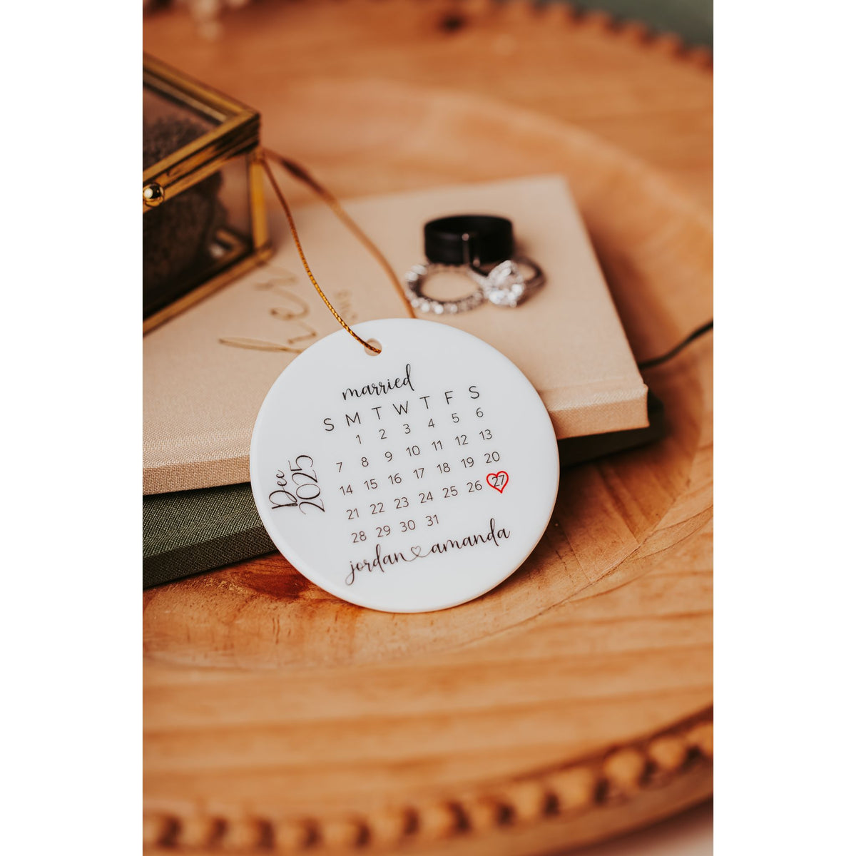The Personalized Wedding Date Ornament – Custom Ceramic Calendar Keepsake by Mirabilia Boutique features “married,” a red heart on December 23rd, and “Jordan & Amanda”—a perfect newlyweds memento beside rings on a wooden tray.