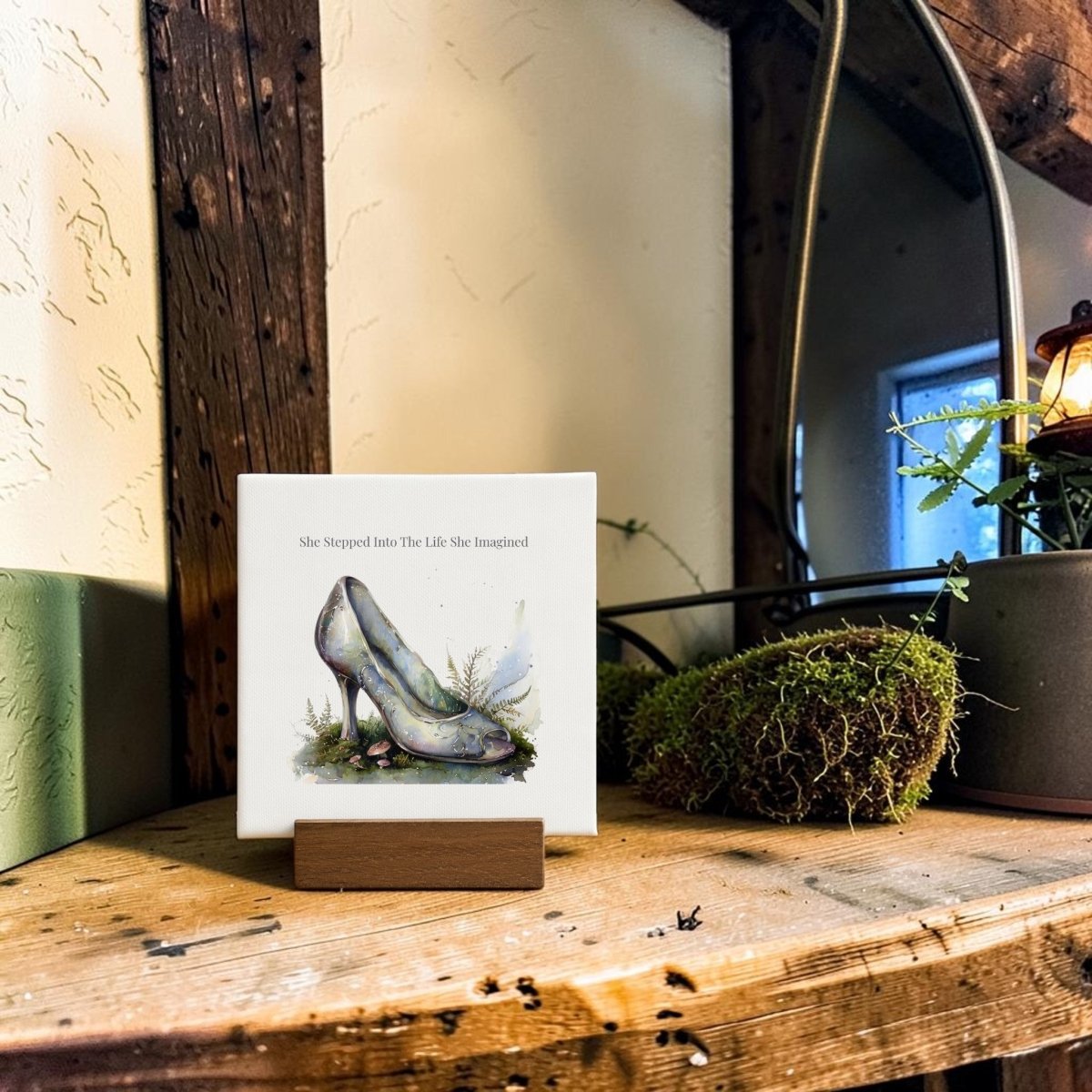 The She Stepped Into the Life She Imagined Mini Sign by Mirabilia Boutique features whimsical glass slipper art and inspiring words displayed on a wooden shelf alongside potted plants, moss, and a rustic lamp—a charming piece of fairytale decor to enliven your space.