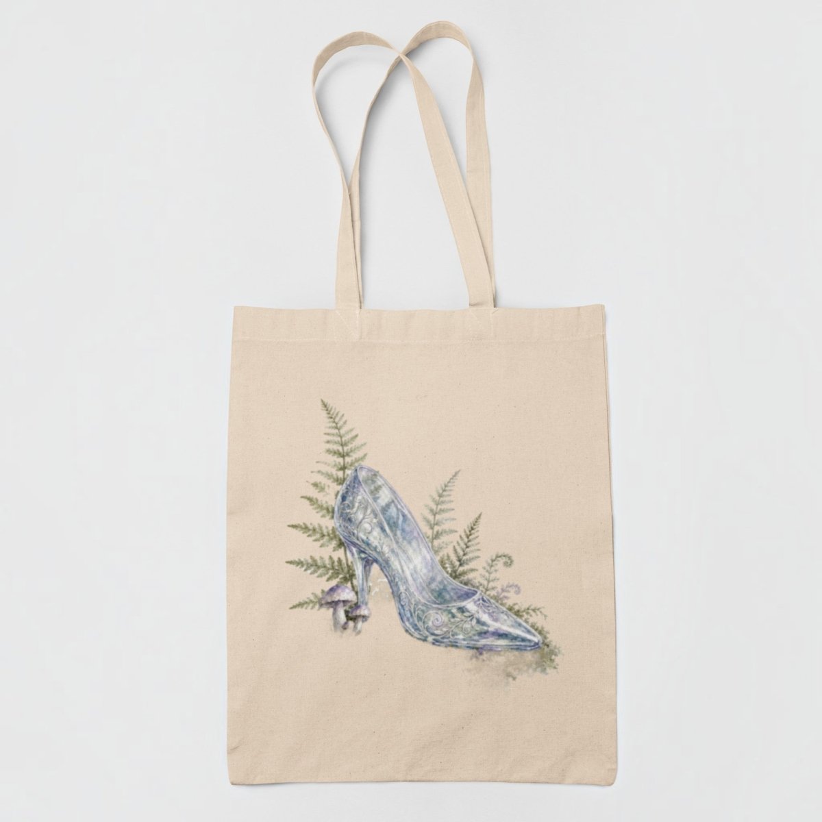 Introducing the Enchanted Glass Slipper Canvas Tote Bag by Mirabilia Boutique—this beige tote features a beautifully centered watercolor illustration of a glass slipper surrounded by green fern leaves, making it the perfect whimsical fairytale tote or charming cottagecore accessory.