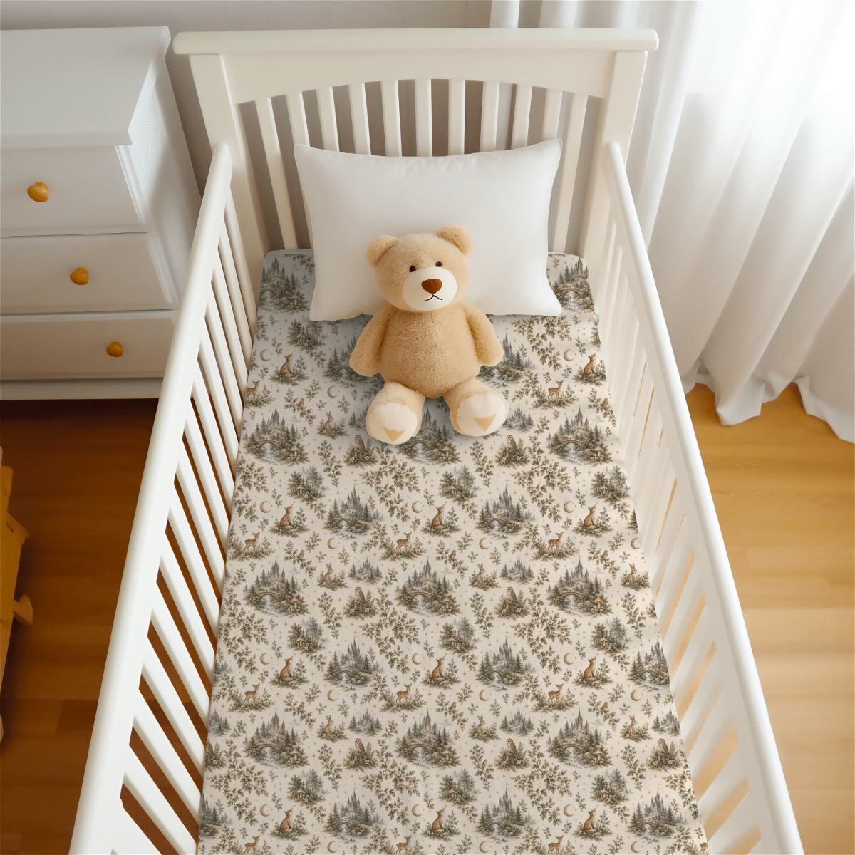 A white crib features the Enchanted Woodlands Crib Sheet by Mirabilia Boutique from the Moon & Moss Nursery collection, accompanied by a white pillow and a light brown teddy bear sitting upright. Nearby stands a white dresser with wooden knobs, while sheer curtains allow natural light to filter in.