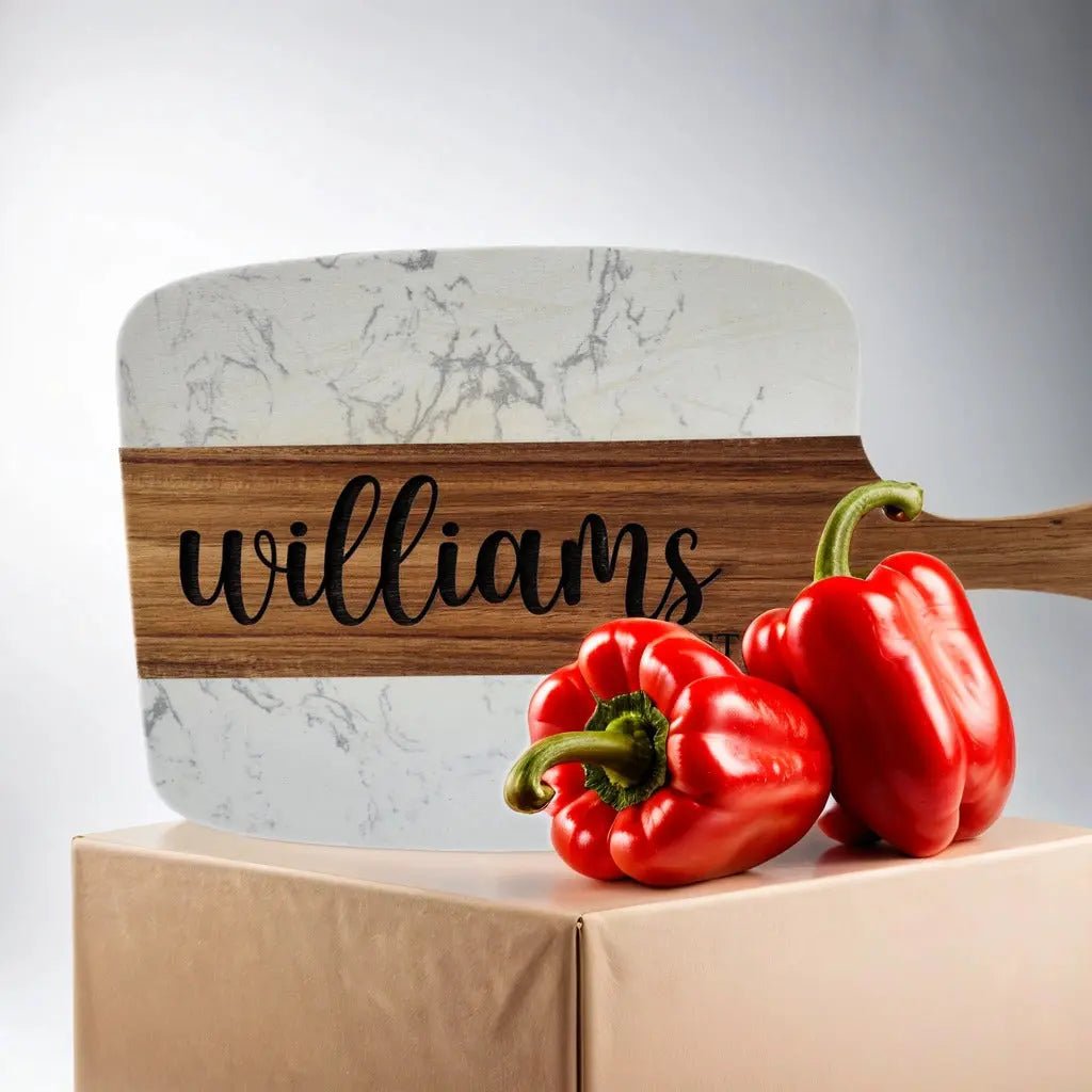 The Personalized Marble and Acacia Serving Board by Mirabilia Boutique, engraved with "williams," is shown behind two red bell peppers on a beige surface.