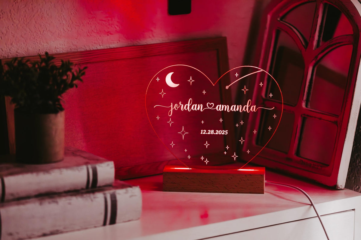 A Mirabilia Boutique Personalized Couples LED Night Light, a heart-shaped acrylic sign with "jordan ♥ amanda," "12.28.2025," stars, and a crescent moon, glows red on a shelf among books and a red window frame.