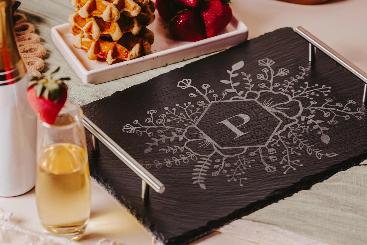 The Personalized Engraved Slate Serving Tray with Handles by Mirabilia Boutique, featuring a floral monogram "P," sits on a table with champagne, waffles, and strawberries—an elegant custom gift idea.