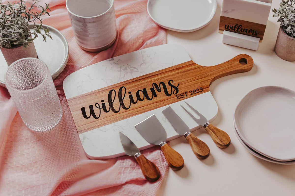The Mirabilia Boutique Personalized Marble and Acacia Serving Board, custom-engraved with a name, creates a stunning charcuterie centerpiece alongside cheese knives, plates, plants, glassware, and a pink textured cloth.