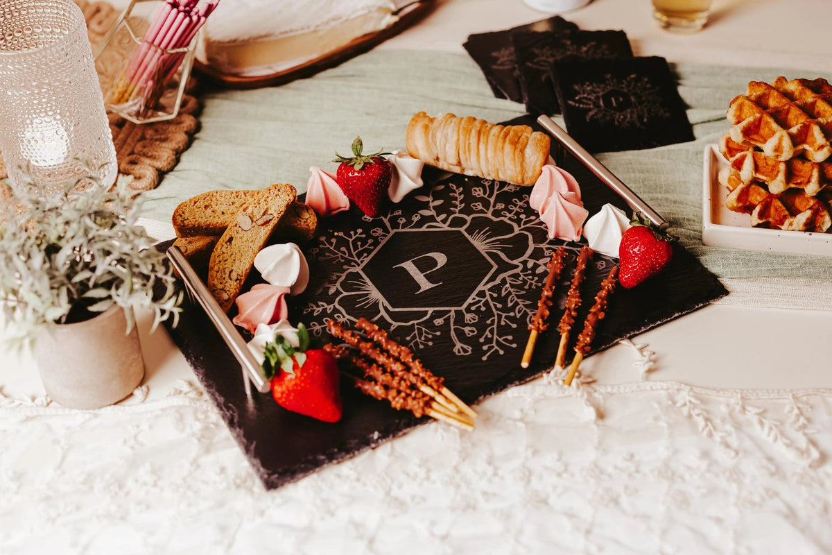 The Mirabilia Boutique Personalized Engraved Slate Serving Tray with Handles, featuring a monogram "P", displays strawberries, breadsticks, biscotti, and meringues. Nearby: a bowl of waffles, napkins, a plant, and candle atop a lace and green tablecloth.