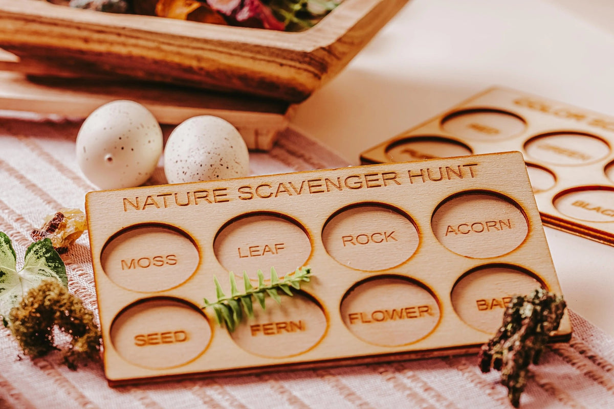 The "Nature Scavenger Hunt for Kids: Fun Outdoor Adventure Game" by Mirabilia Boutique features a wooden board with labeled slots for gathering moss, leaf, rock, acorn, seed, fern, flower, and bark. This eco-friendly outdoor toy is displayed on a table surrounded by speckled eggs and other small nature treasures.