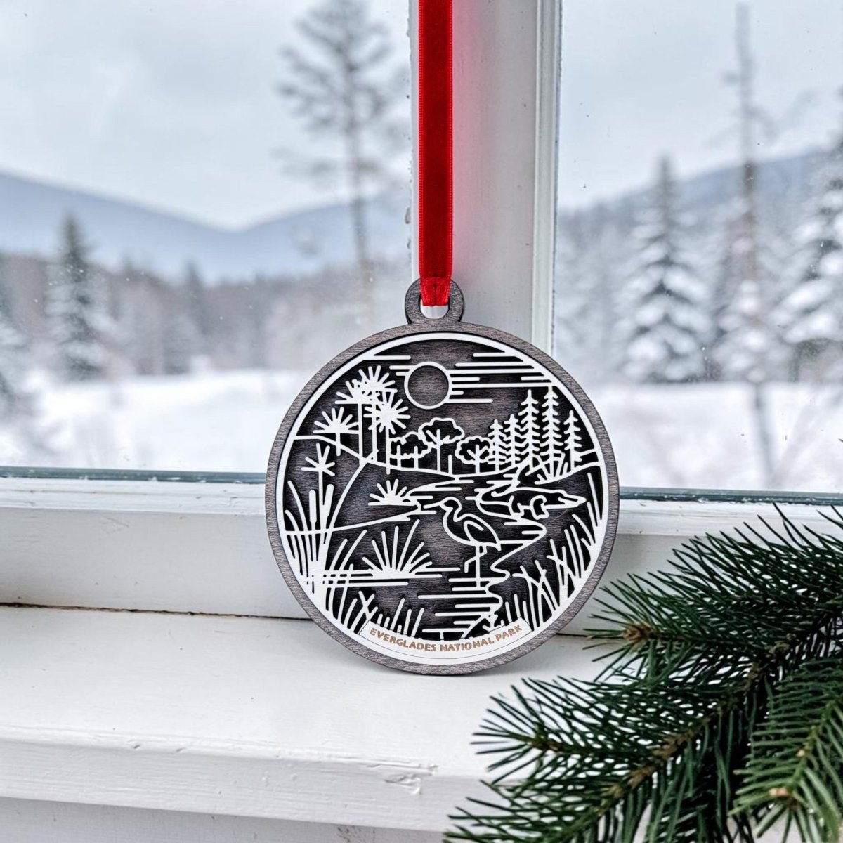 A Mirabilia Boutique Everglades National Park Line Art Christmas Ornament, featuring an engraved silver nature scene, hangs by a red ribbon in front of a snowy window while evergreen branches accent the corner.