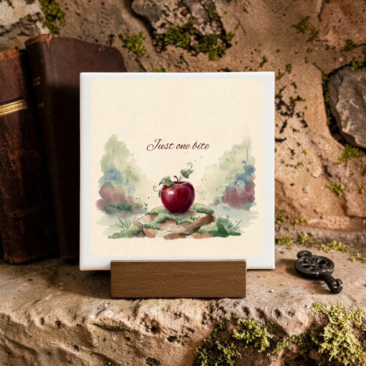 The Just One Bite Mini Canvas Sign by Mirabilia Boutique features a red apple and the phrase "Just one bite" on a watercolor background. Displayed on a wooden stand, this small canvas sits atop a stone surface surrounded by vintage books, moss, and an ornate key—adding the perfect woodland charm to your fairy tale decor.