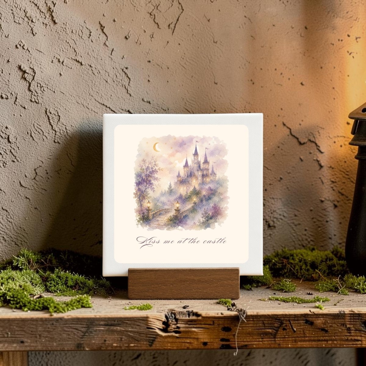 The Kiss Me at the Castle Mini Canvas Sign by Mirabilia Boutique features a dreamy castle scene with trees and a crescent moon, displayed on a wooden stand. Below the enchanting artwork is the phrase “Kiss me at the castle.” Accented with moss and soft lighting, this romantic home decor piece makes a perfect gift or accent for anyone who loves fairytale-inspired style.