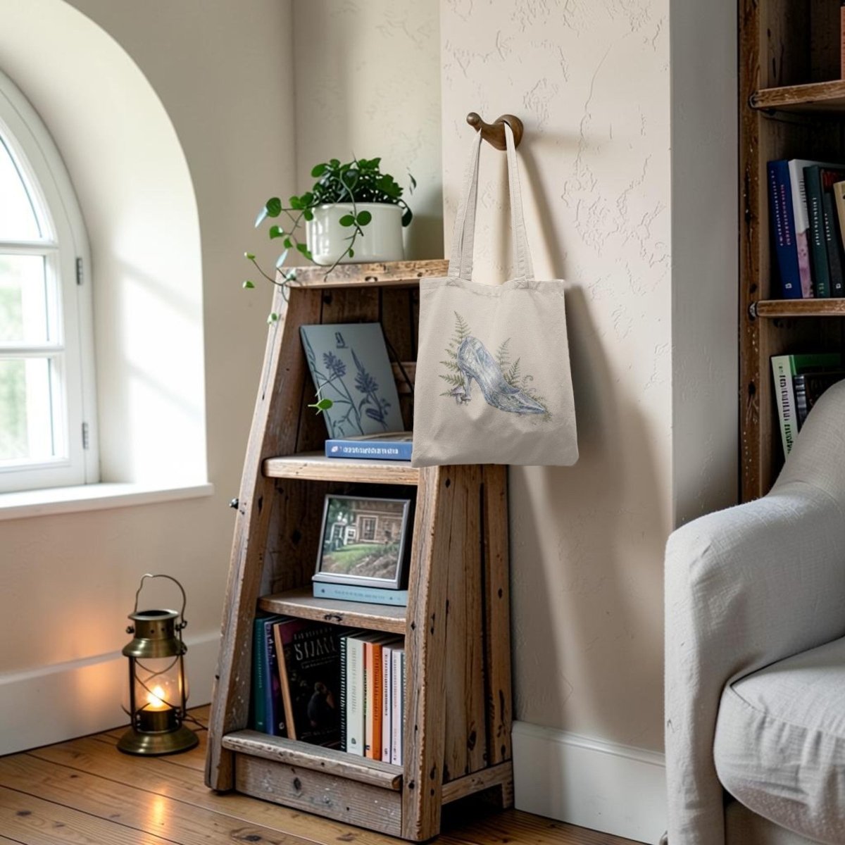 A rustic wooden bookshelf holds books, a framed photo, a potted plant, and the Enchanted Glass Slipper Canvas Tote Bag by Mirabilia Boutique, featuring a charming blue bird design. A lantern rests on the floor while sunlight streams in through an arched window.