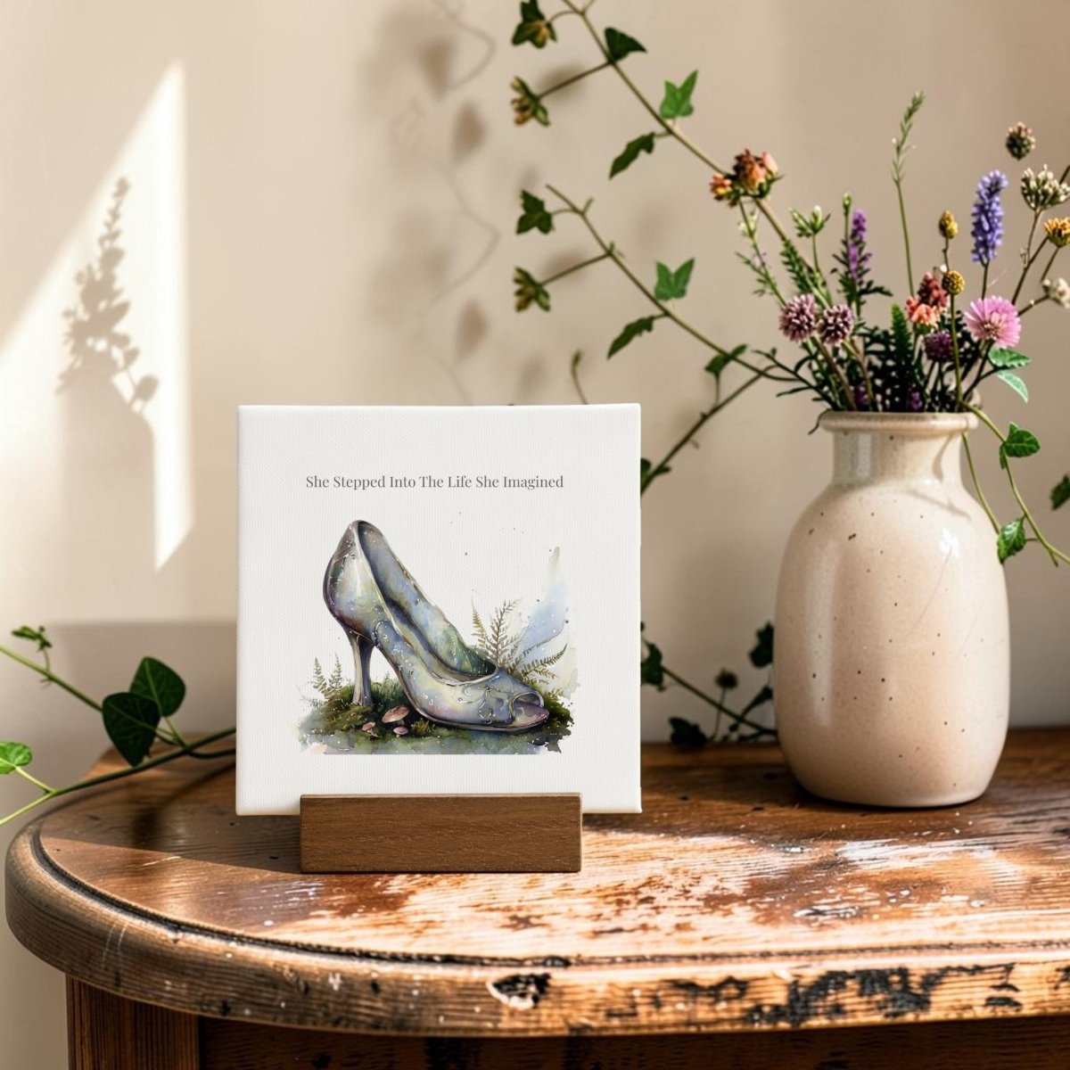 The She Stepped Into the Life She Imagined Mini Sign from Mirabilia Boutique showcases painted high heels inspired by glass slipper art and displays the uplifting phrase, "She Stepped Into The Life She Imagined." Placed on a wooden stand beside a beige vase of wildflowers on a rustic wooden table, it creates the perfect fairytale-inspired decor.