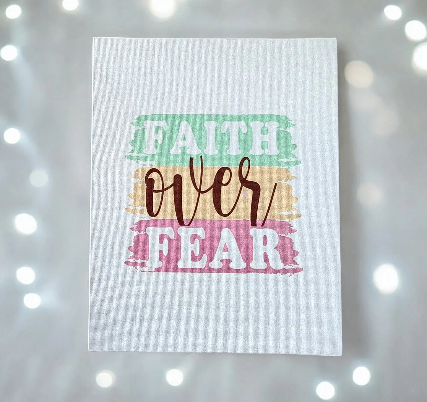 Presenting the "Faith Over Fear" Canvas Art by Mirabilia Boutique, a piece of inspirational wall decor for your home. This motivational artwork showcases "Faith Over Fear" prominently on a pastel-themed background with teal, peach, and pink paint-like stripes. The canvas features softly blurred effects complemented by glowing lights to provide an inspiring touch.