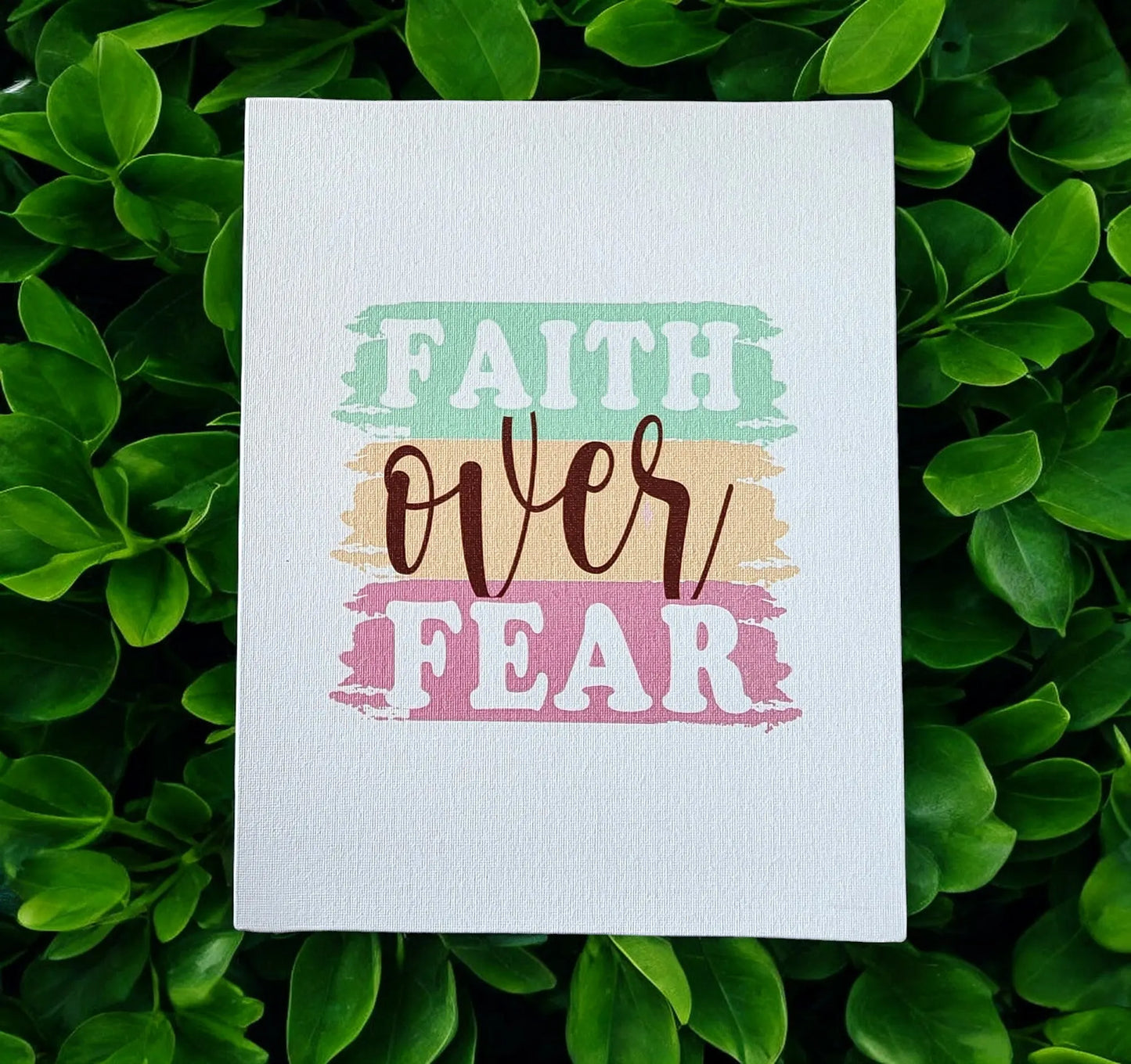 The Faith Over Fear Canvas Art from Mirabilia Boutique features the motivational phrase "Faith Over Fear" beautifully accentuated with pastel shades of green, orange, and pink. With its lush green leaf backdrop, this inspirational wall decor makes an ideal gift for adding a serene and uplifting ambiance to any home.
