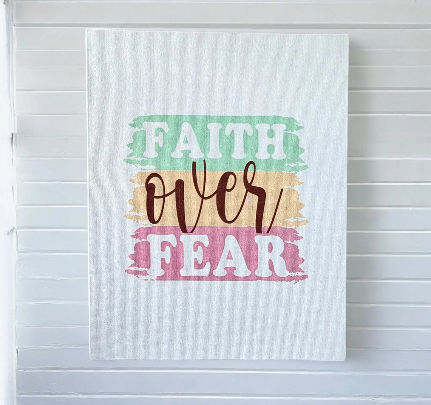 A piece from Mirabilia Boutique, the Faith Over Fear Canvas Art, adorns a white wall with its motivational design. The artwork displays the phrase "Faith Over Fear" in vibrant, stylistic text; the bold pastels of "Faith" and "Fear" stand out against the graceful cursive of "Over," making it an ideal inspiring addition to any home decor.