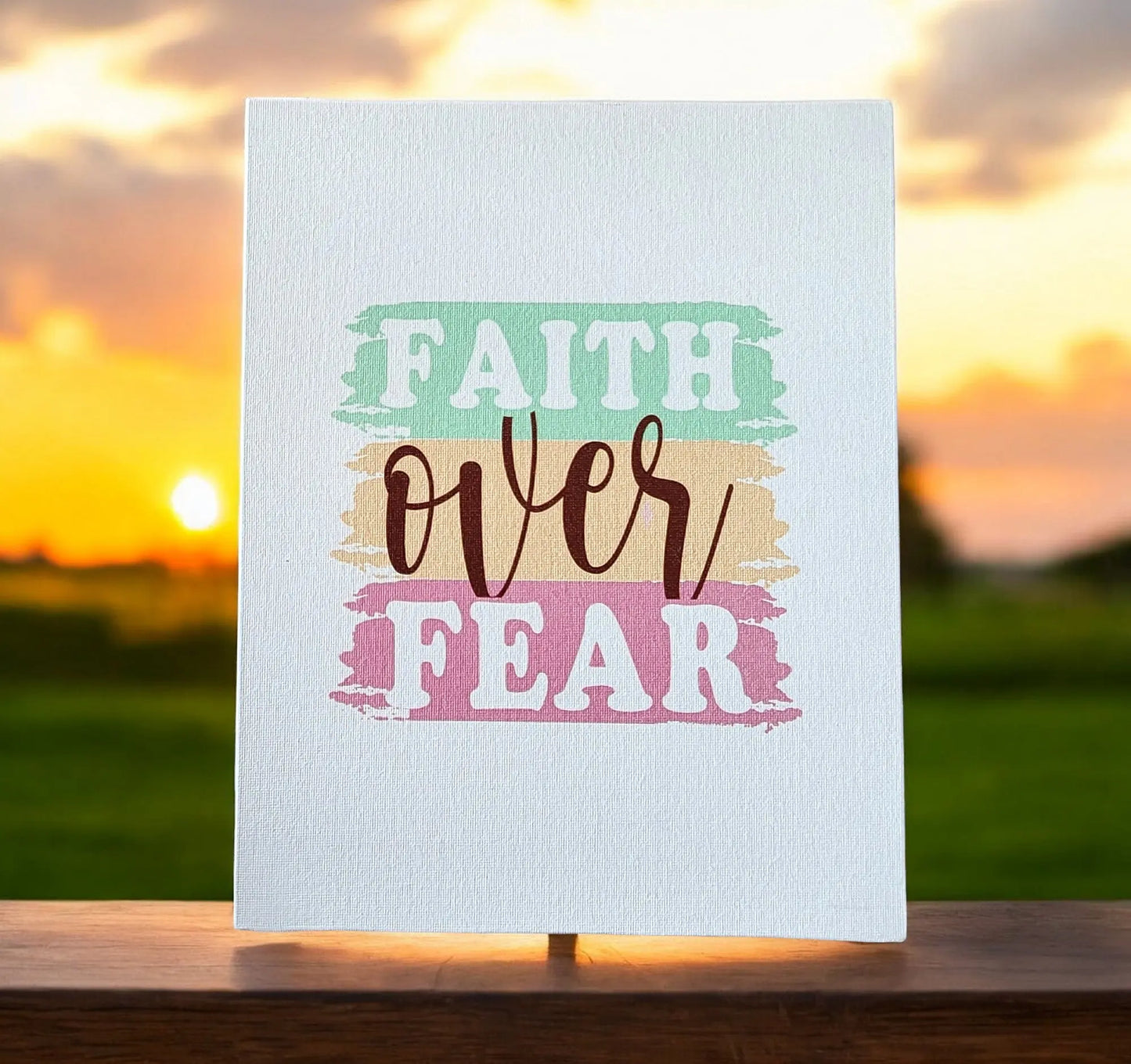 This inspiring gift from Mirabilia Boutique features the "Faith Over Fear" canvas art, displayed on an easel with its vibrant typography set against a blurred sunset backdrop in shades of green, orange, pink, and brown.