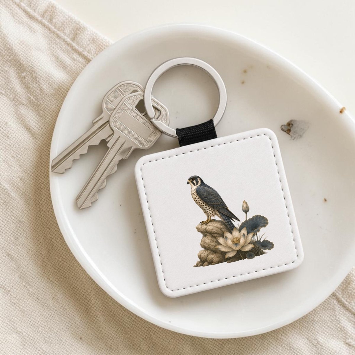 The Falcon & Lotus Keychain | Ancient Egyptian Art by Mirabilia Boutique—a white keychain featuring a falcon and lotus inspired by ancient Egyptian art—rests on a dish beside two silver keys.