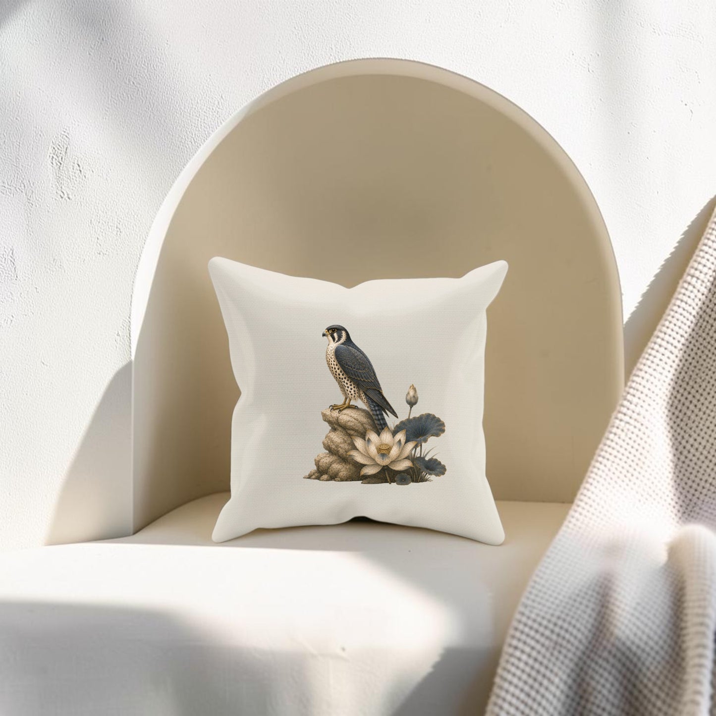 A Falcon & Lotus Pillow by Mirabilia Boutique, featuring an illustration of a falcon perched among rocks and lotus flowers, accents a cream-colored chair bathed in soft sunlight, with a textured blanket nearby.