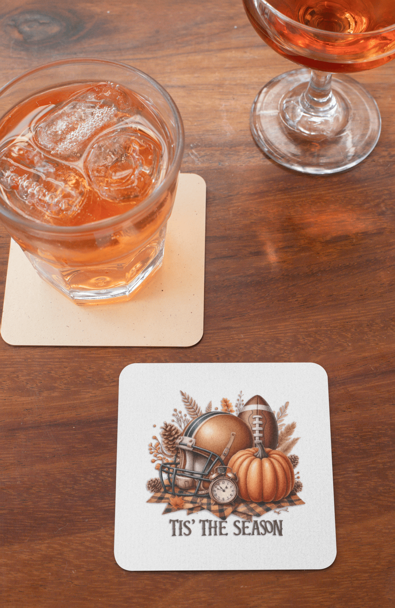 A glass of iced drink and a cocktail rest on a wooden table with Mirabilia Boutique’s “Tis’ the Season Coaster Set” featuring football, pumpkin, and autumn designs—perfect fall decor or gift for game day.