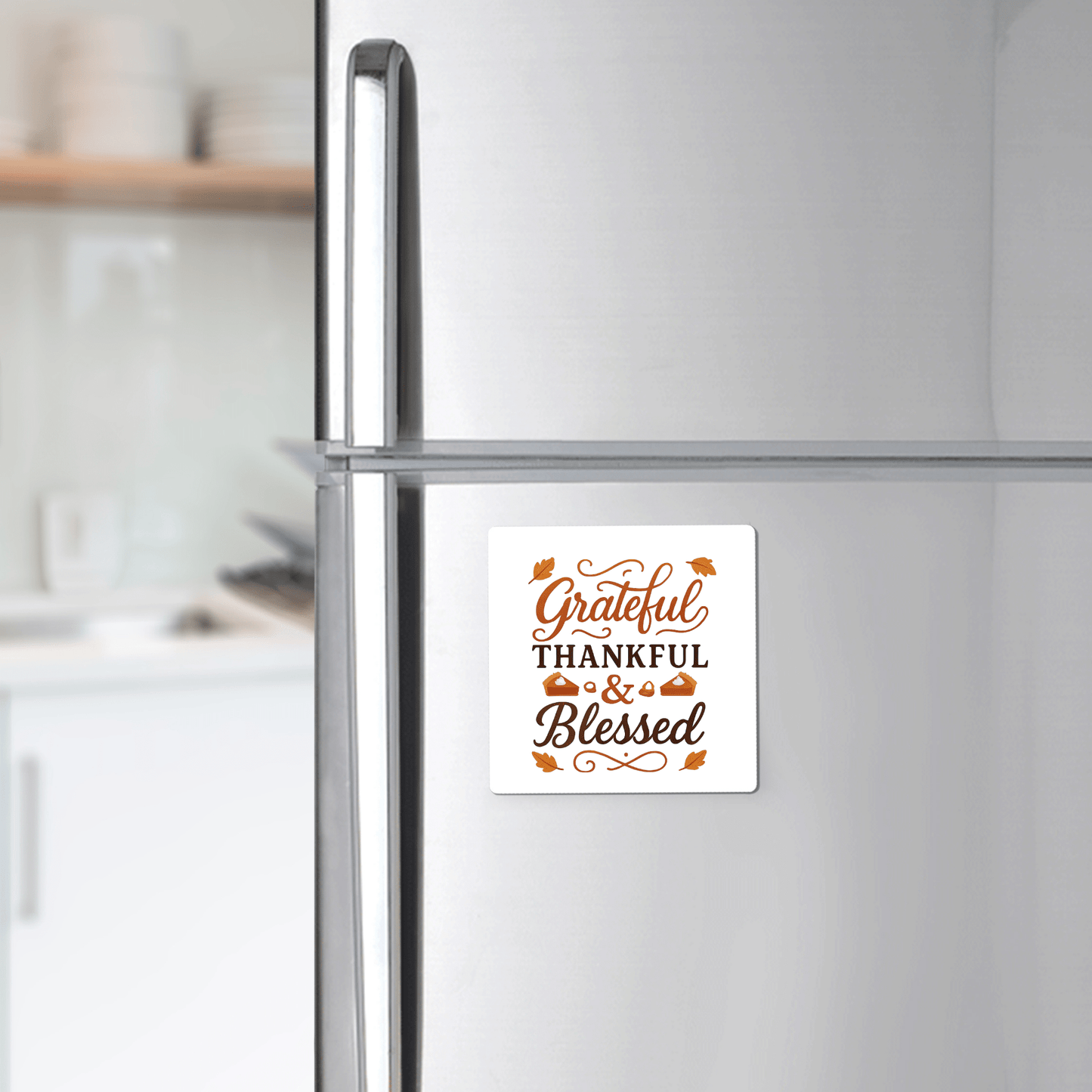 A silver fridge displays the Mirabilia Boutique "Grateful Thankful Blessed Magnet" in brown and orange with leafy accents—an ideal fall or Thanksgiving decor piece. A blurred kitchen is visible in the background.