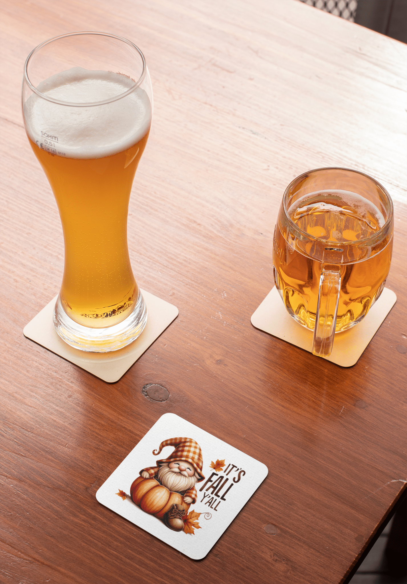 A glass of beer and a mug of cider rest on Mirabilia Boutique’s Fall Gnome Coasters Set, featuring pumpkins, leaves, and "It's Fall Y'all"—perfect drink decor to add charm to your autumn home.