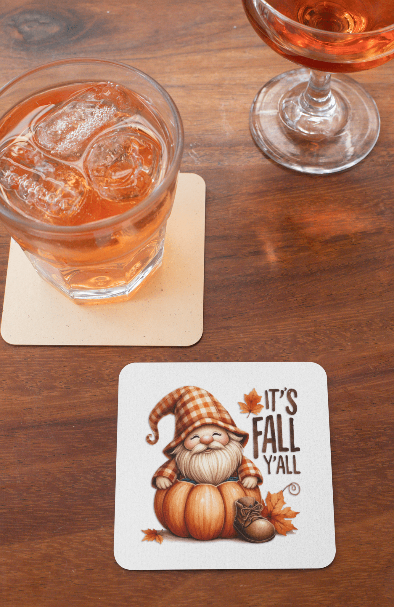 A drink with ice and a glass of rosé rest on Mirabilia Boutique’s Fall Gnome Coasters Set featuring a gnome in a plaid hat on a pumpkin with autumn leaves and "IT'S FALL Y'ALL" text. Perfect seasonal drink decor!.