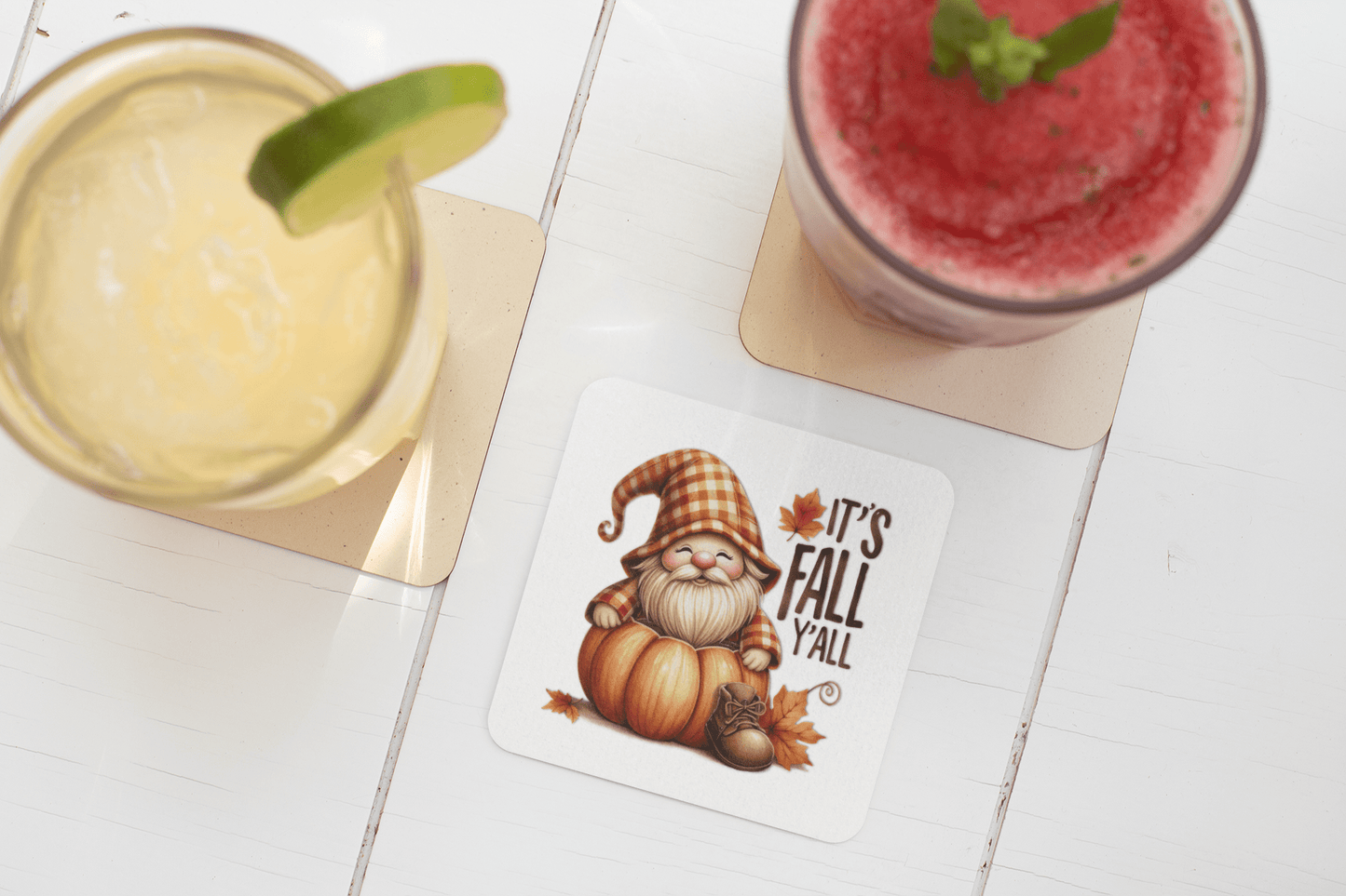 Mirabilia Boutique's Fall Gnome Coasters Set features a gnome in a striped hat with pumpkins, leaves, and "It's Fall Y'all"—a charming drink decor piece for your autumn home. Shown beside two drinks on a white table.