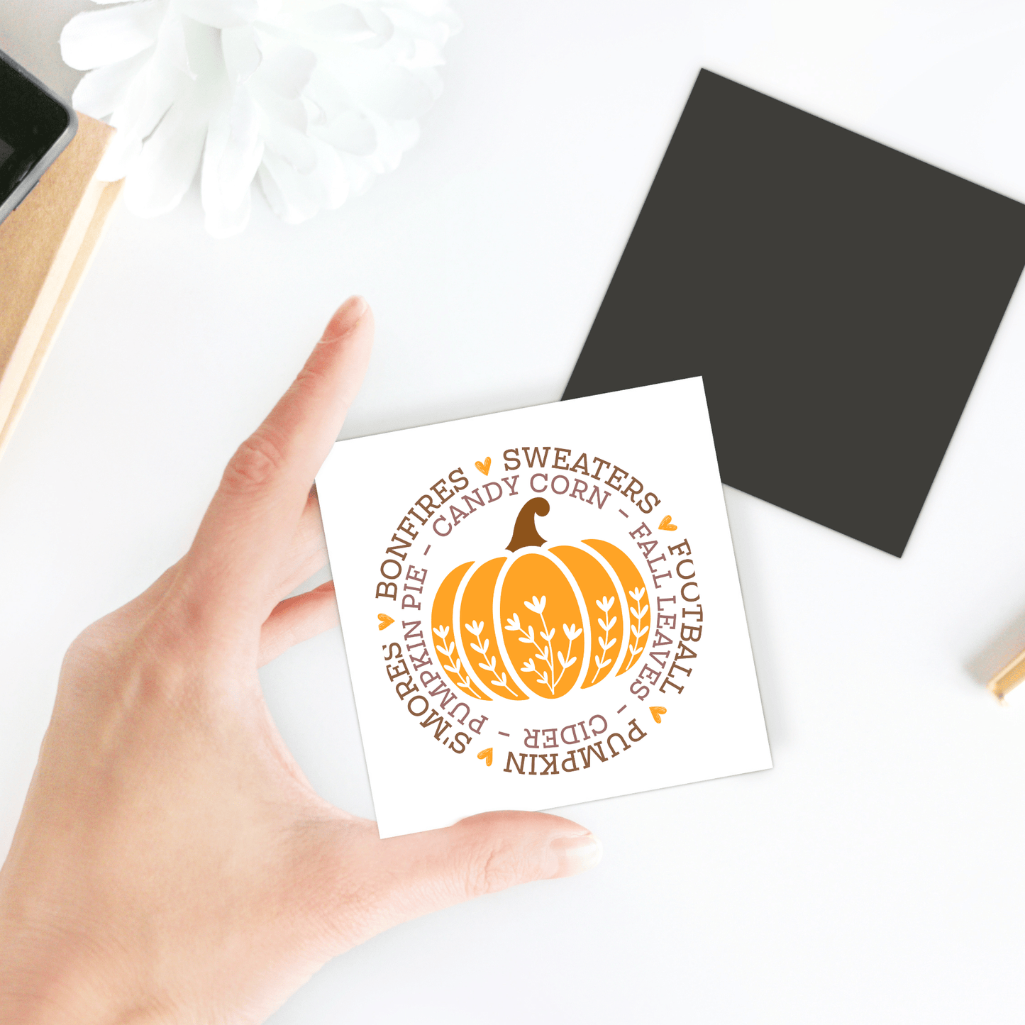 A hand displays the Fall Pumpkin Magnet by Mirabilia Boutique, featuring an orange pumpkin and autumn words like "sweaters" and "cider"—a perfect seasonal fridge art or Thanksgiving gift for your autumn kitchen decor.