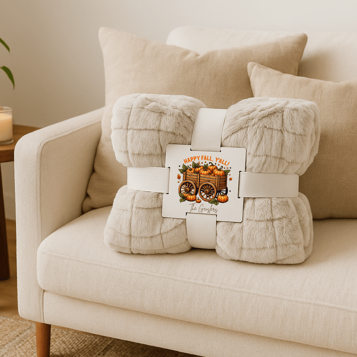 A beige sofa with cream pillows showcases Mirabilia Boutique's Custom Fall Throw Blanket, featuring a festive tag with pumpkins, sunflowers, and “Happy Fall Y’all!”—a perfect cozy autumn gift for the home. A candle glows on a side table nearby.