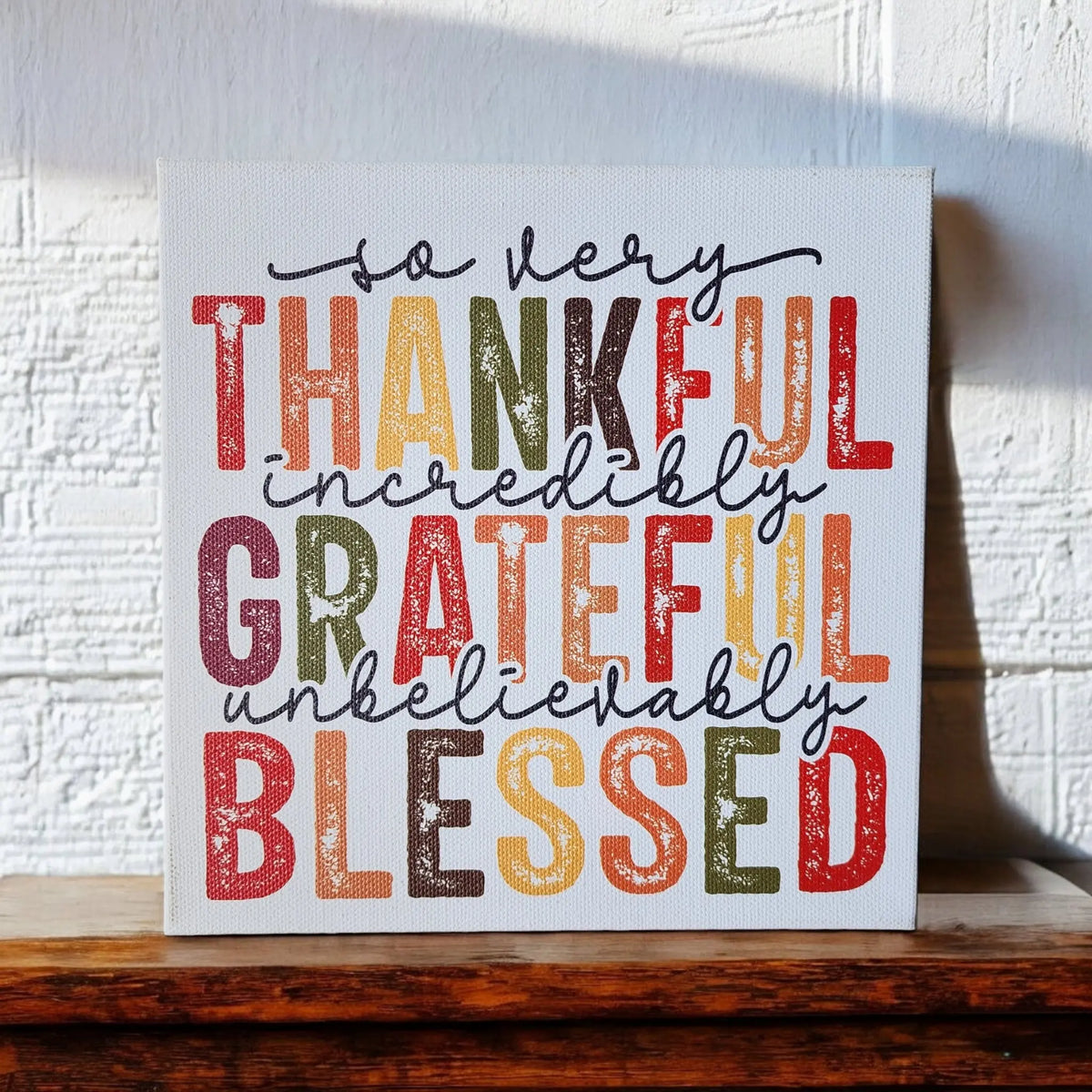 A canvas from Mirabilia Boutique, featuring colorful and bold text, is propped against a white textured wall. The "Thankful Grateful Blessed Inspirational Sign for Seasonal Home Decor" reads: "so very Thankful, incredibly Grateful, unbelievably Blessed." Embracing Thanksgiving colors—shades of red, orange, yellow, green, and brown—this home decor sign is perfect for any cozy space. The canvas rests gracefully on a wooden surface.