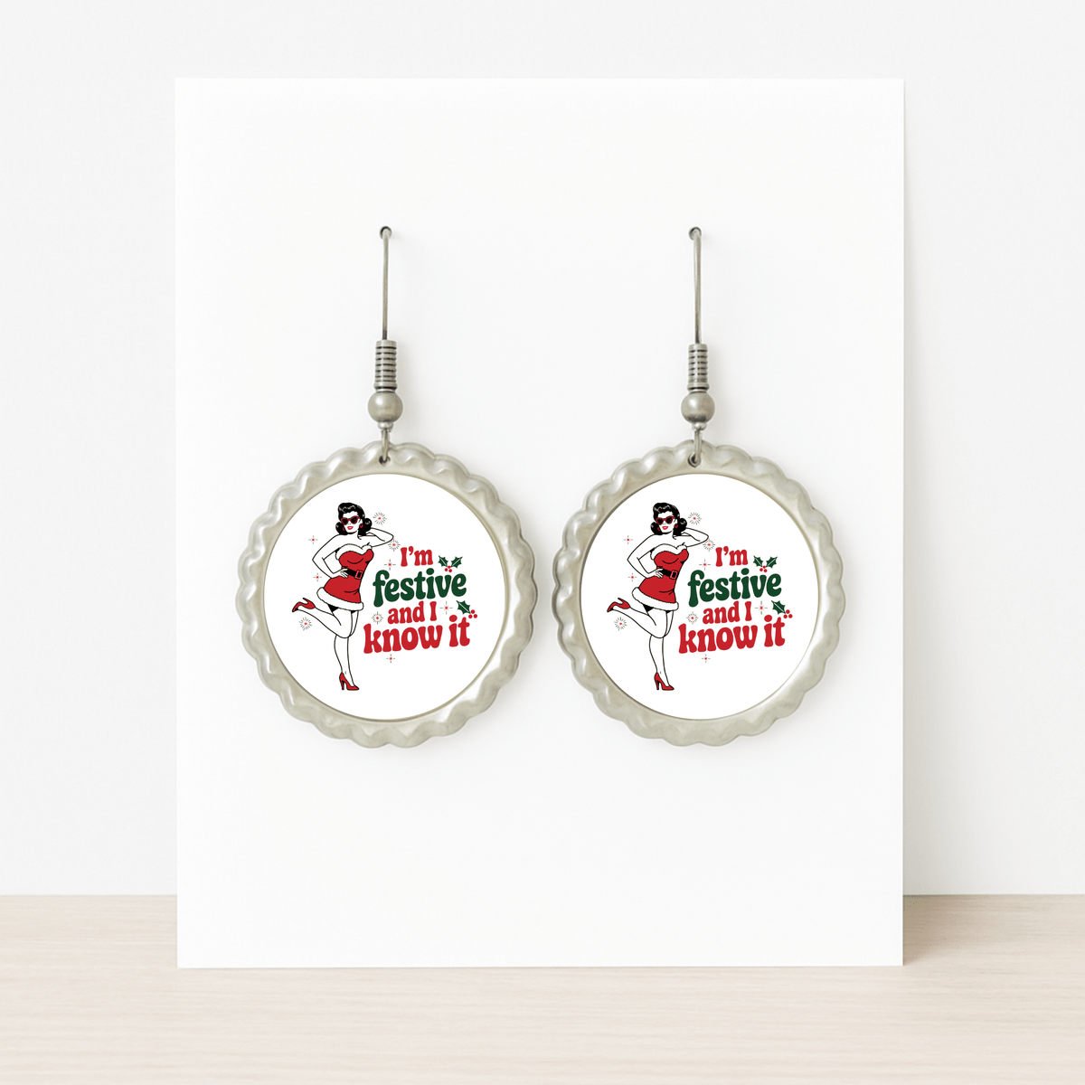 The Festive and I Know It Earrings by Mirabilia Boutique feature a retro pin-up girl in a Santa hat and red dress with "I'm festive and I know it" text, displayed on a white card—perfect for holiday flair.