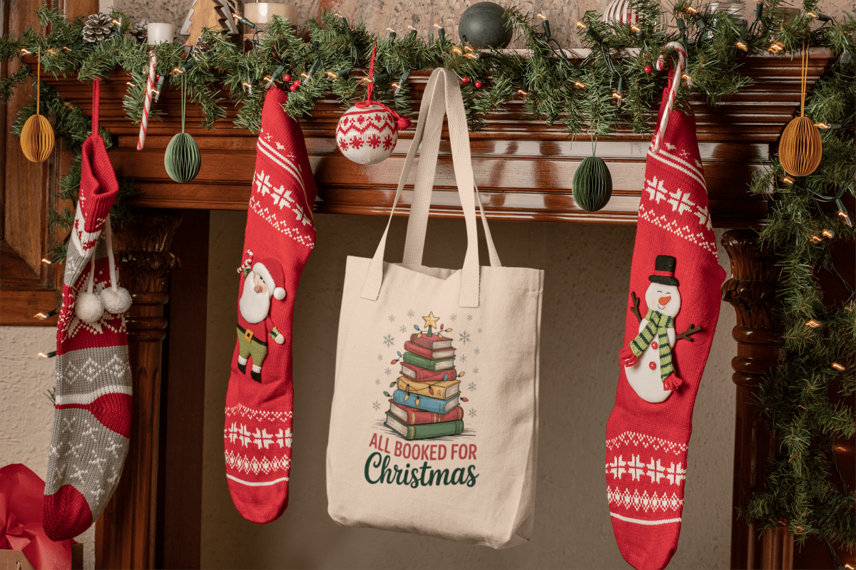 A festive mantel with garland, ornaments, three red stockings, and the Mirabilia Boutique "All Booked for Christmas Book Stack Tote Bag"—a perfect gift for book lovers.