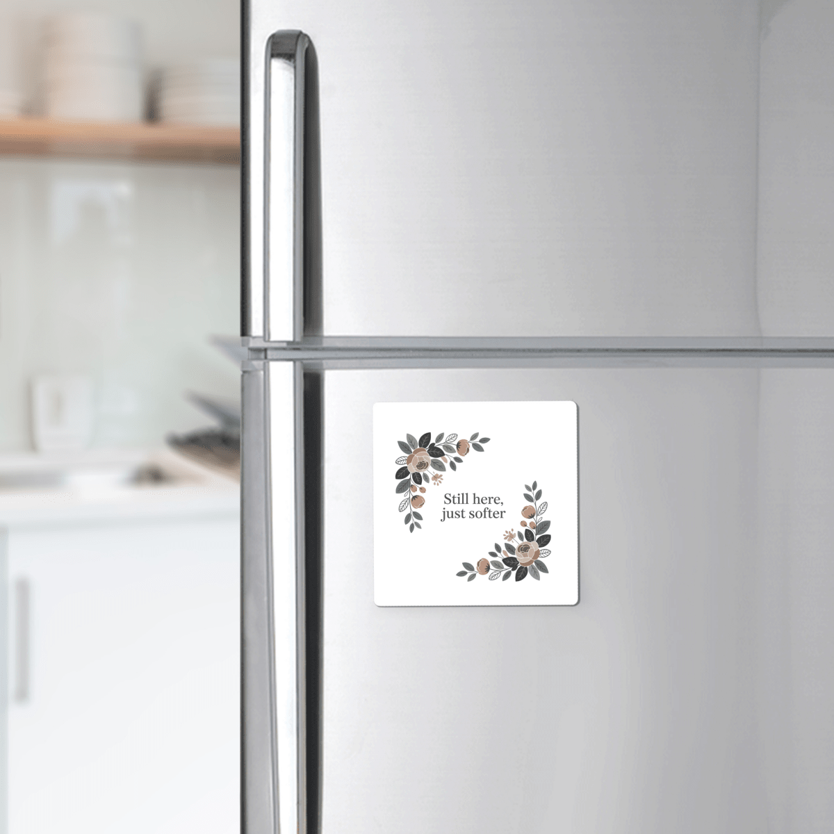 A silver fridge displays the "Still Here Just Softer Floral Magnet" by Mirabilia Boutique, featuring a botanical design and gentle message—an ideal encouragement gift for any softly blurred kitchen scene.