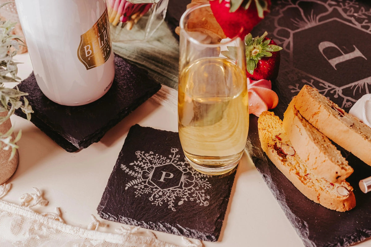 A glass of white wine sits on a Mirabilia Boutique Personalized Slate Coaster, engraved with a floral monogram "P," alongside treats—an elegant, custom housewarming gift from the Engraved Monogram Set of 4.