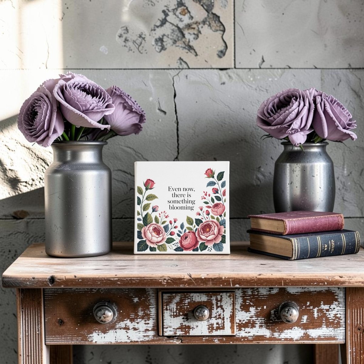 The Even Now There Is Something Blooming Canvas Wall Art by Mirabilia Boutique shows a rustic table with silver vases, purple flowers, stacked books, and an inspiring card—perfect as an encouraging gift or floral wall decor.