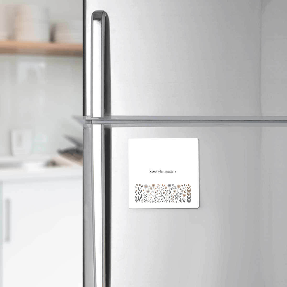 A Mirabilia Boutique Keep What Matters Floral Magnet, featuring an inspiring quote and flower illustration, adds a charming touch to any stainless steel fridge—ideal for gifting or enhancing cottage décor. A blurred kitchen is seen in the background.