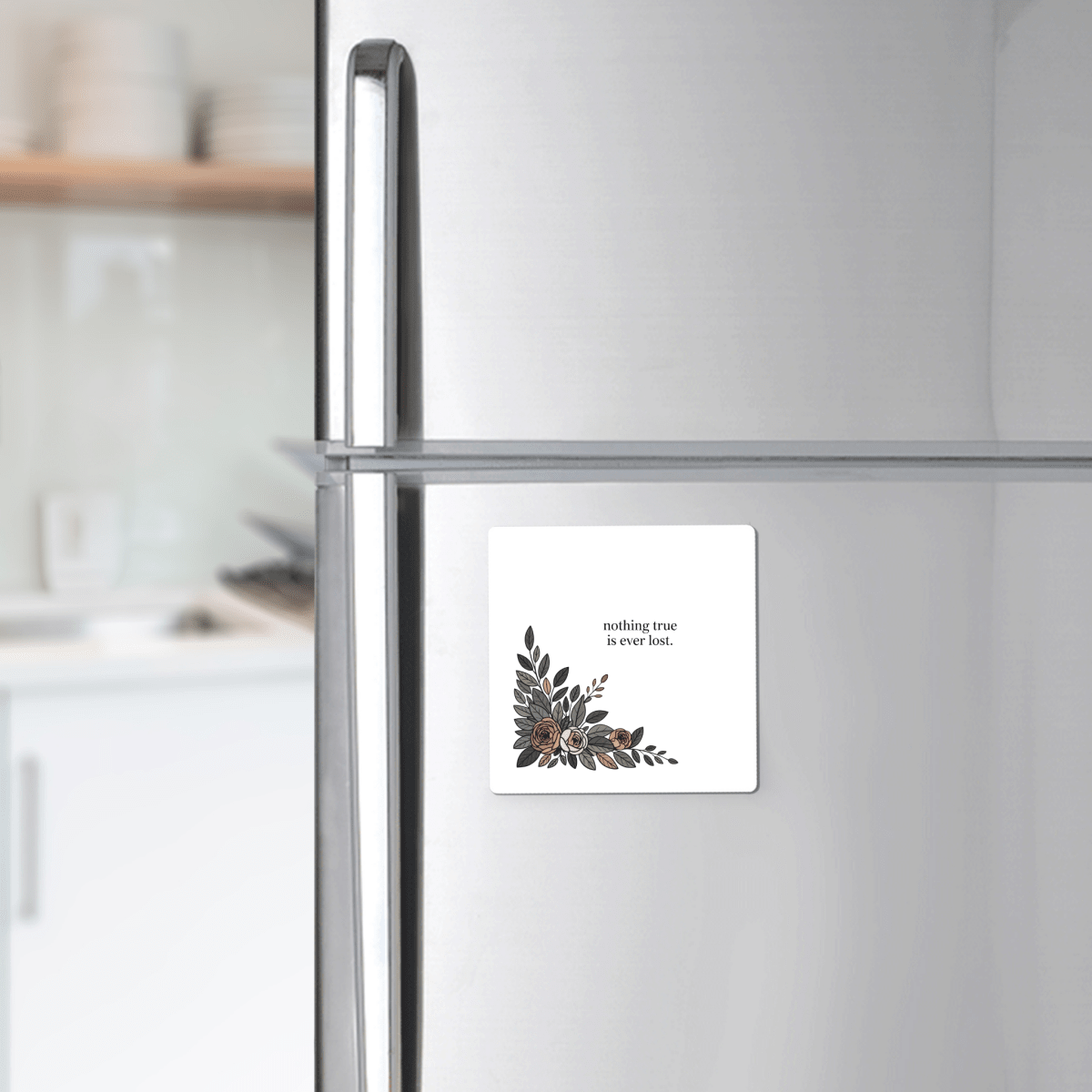 A silver refrigerator holds the Mirabilia Boutique "Nothing True Is Ever Lost Floral Magnet," featuring delicate corner artwork. Ideal as an encouragement gift, it stands out with dishes on a kitchen counter in the background.