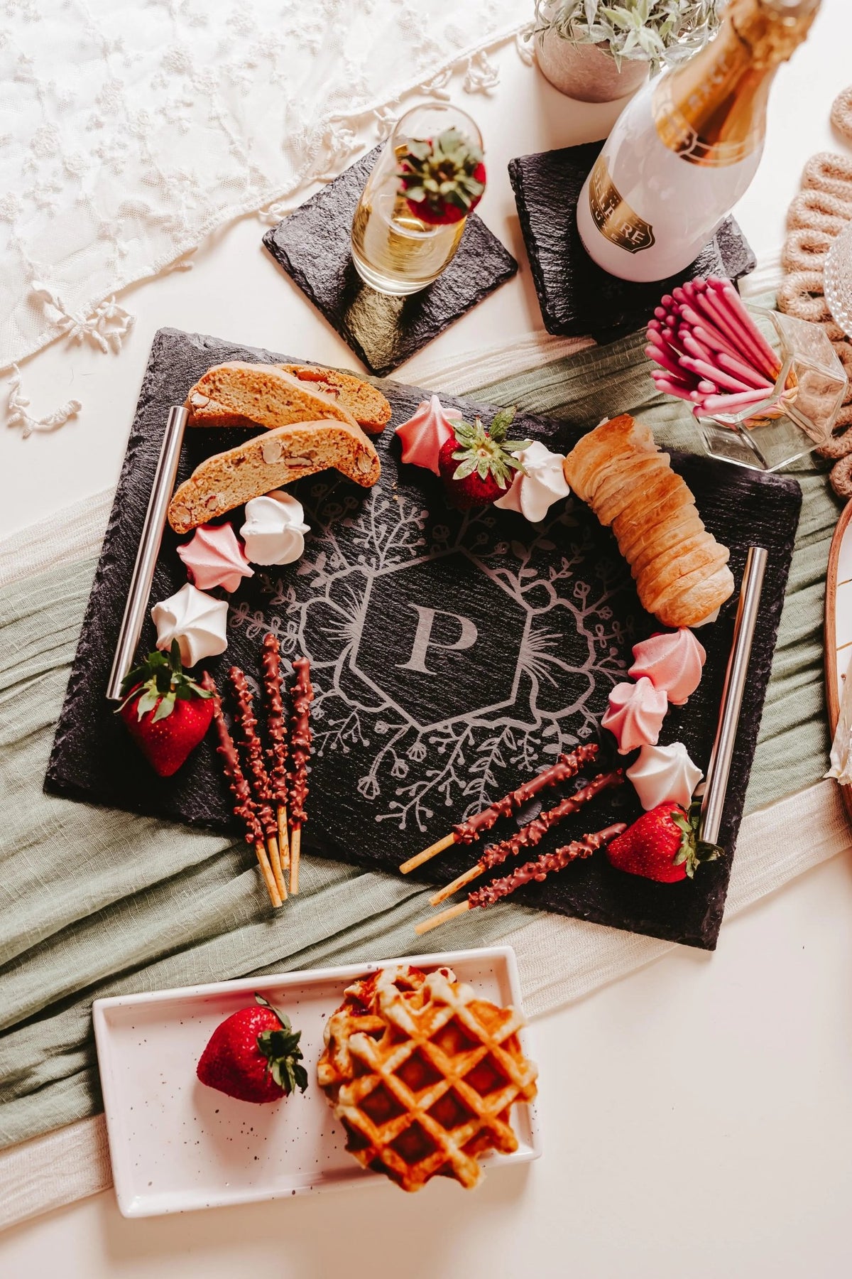 The Mirabilia Boutique Personalized Engraved Slate Serving Tray with Handles, featuring a floral monogram “P”, is elegantly displayed with strawberries, meringues, biscotti, chocolate sticks, a croissant, and a sparkling drink—perfect for gifting or entertaining.