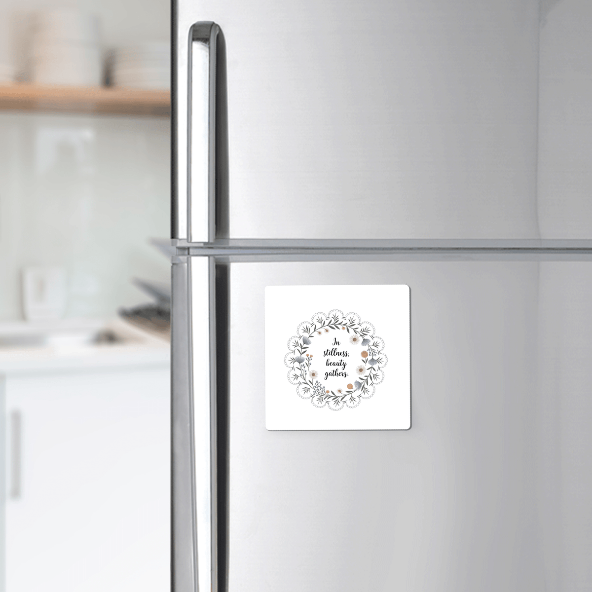 A silver refrigerator displays Mirabilia Boutique's "In Stillness Beauty Gathers" floral quote magnet, featuring a wreath design. A white kitchen with shelves and dishes appears softly blurred in the background, creating mindful decor.