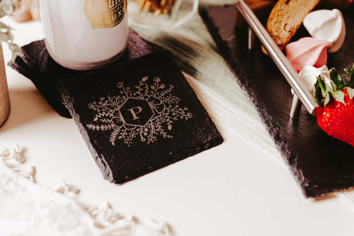 The Personalized Slate Coasters – Engraved Monogram Set of 4 by Mirabilia Boutique features a black coaster with a floral "P" design, shown on a white surface near treats—a perfect custom housewarming gift.