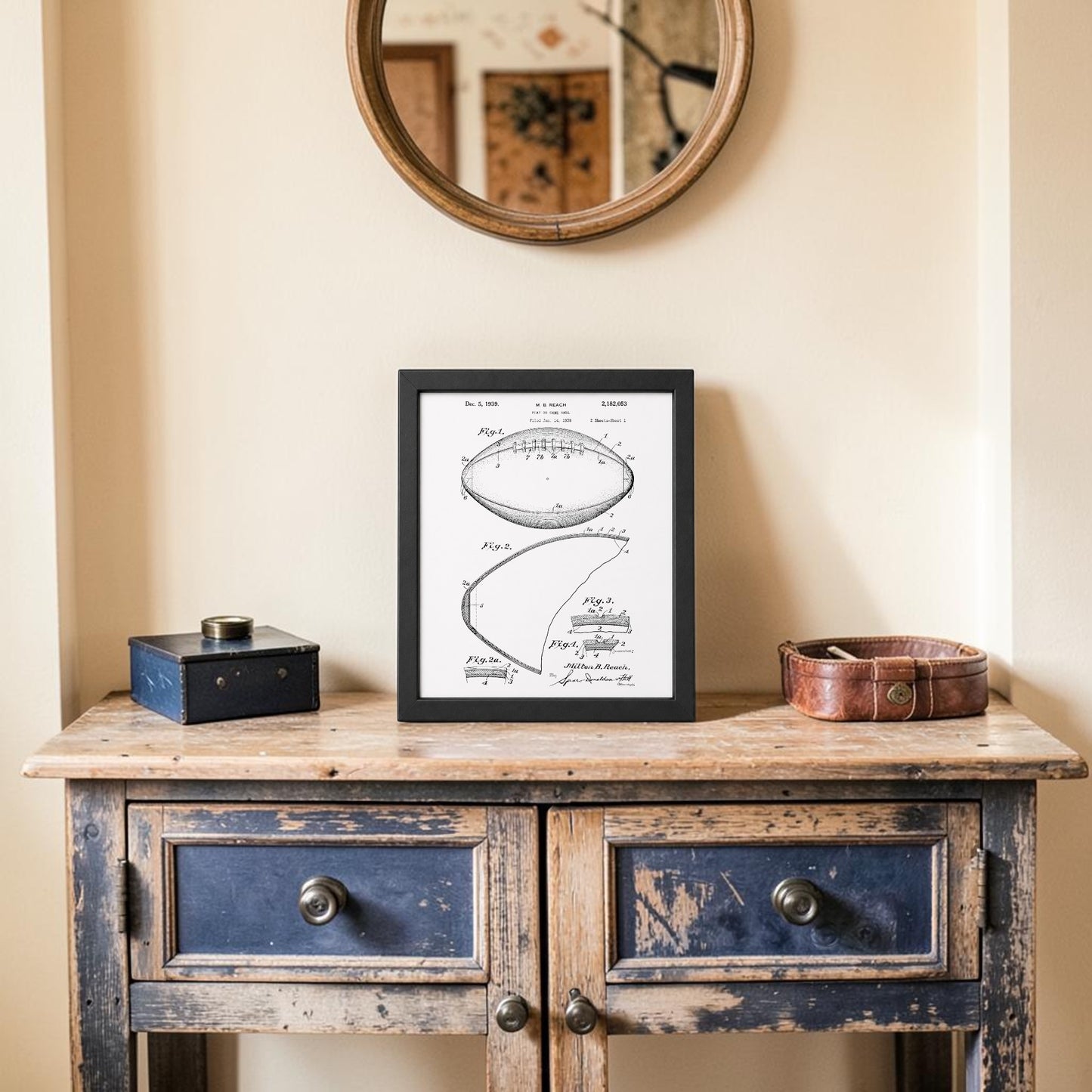 A rustic wooden table with two drawers displays the Mirabilia Boutique American Football Patent Art Print 8x10 in a frame, a round leather tray, and a small blue box. Above, a round mirror hangs on the cream-colored wall.