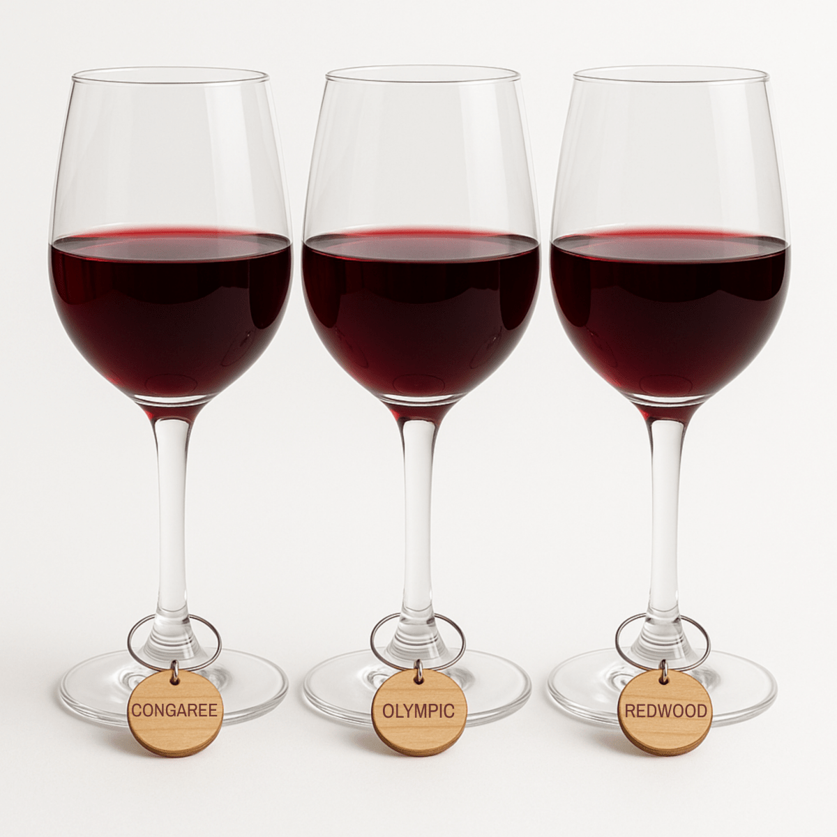 Three glasses of red wine, each with a Mirabilia Boutique Forest And Fog Wine Charm labeled "CONGAREE," "OLYMPIC," or "REDWOOD," arranged in a row—ideal for nature lovers who cherish forest scenery.
