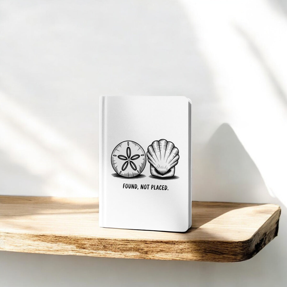 The Found, Not Placed Journal – Coastal Quote Notebook by Mirabilia Boutique, featuring white seashell designs and the phrase "Found, not placed.", rests on a sunlit wooden shelf casting soft shadows against a white wall.