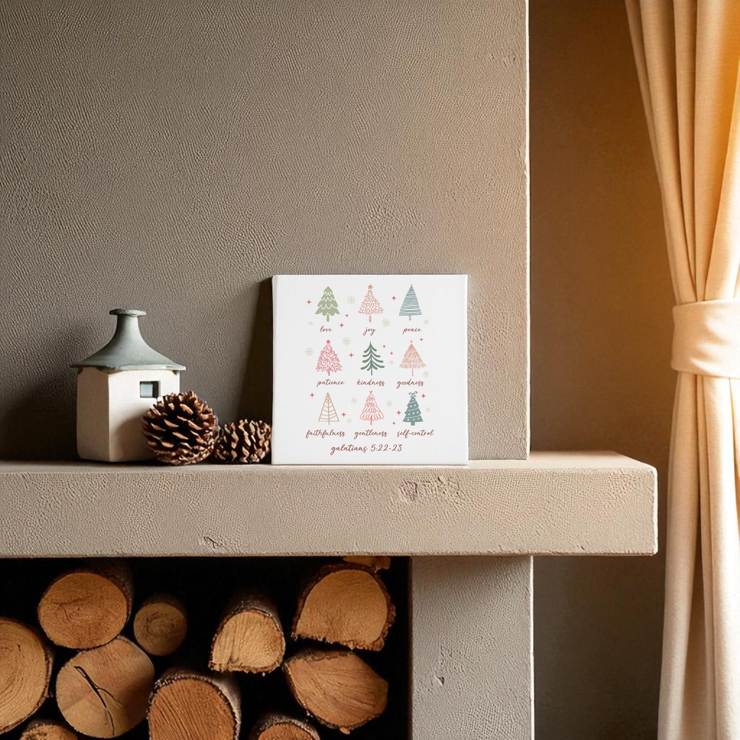 A decorated mantel displays a small ceramic house, pine cones, and the Mirabilia Boutique 'Fruits of the Spirit' Christian Christmas Canvas featuring pastel trees and Galatians 5:22. Firewood is neatly stacked below, near a beige curtain.