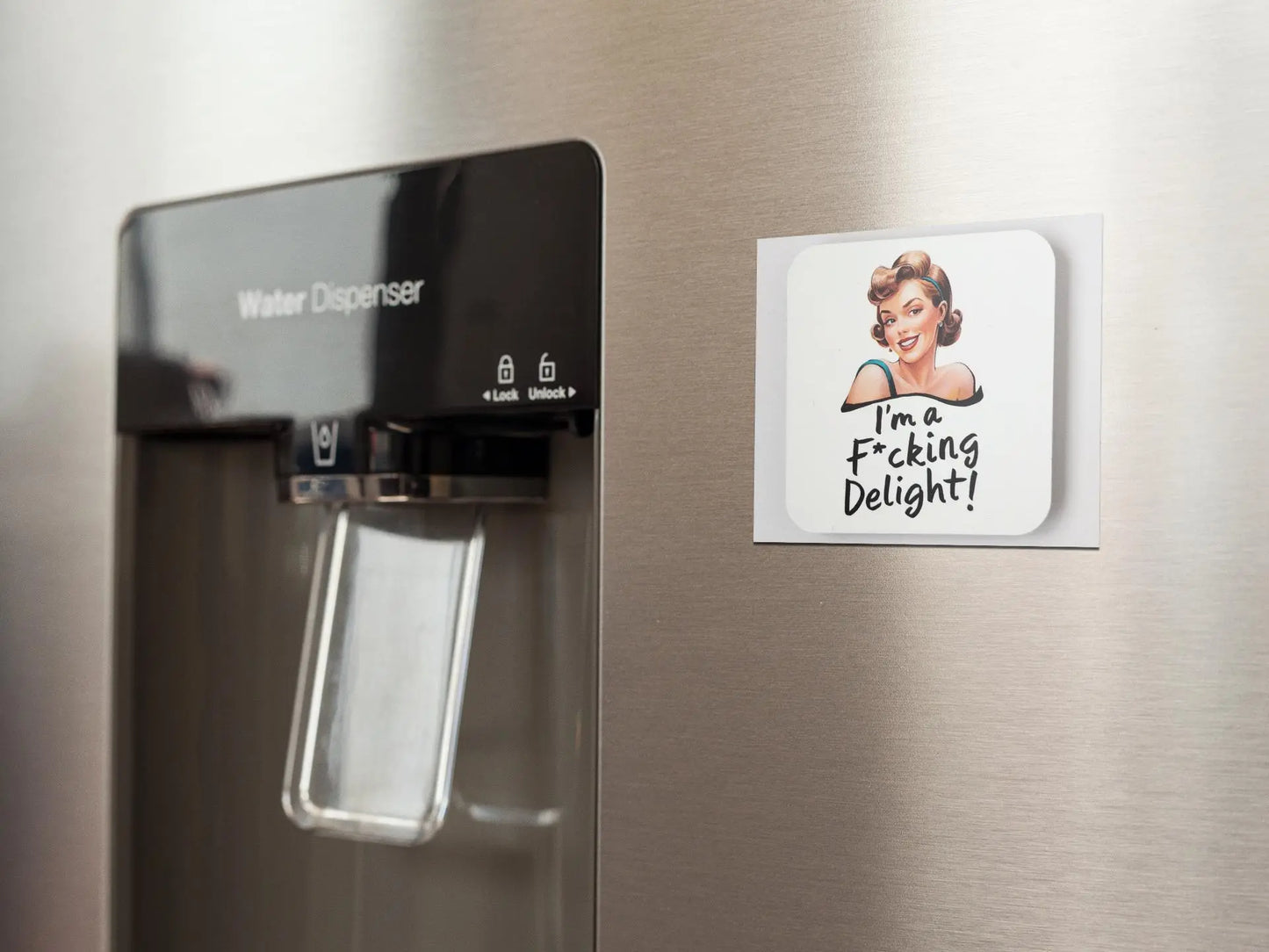 A close-up image of a water dispenser on a stainless steel refrigerator, next to a snarky retro magnet from Mirabilia Boutique featuring a cartoon of a woman. The humorous fridge magnet reads, "I'm a Fucking Delight!