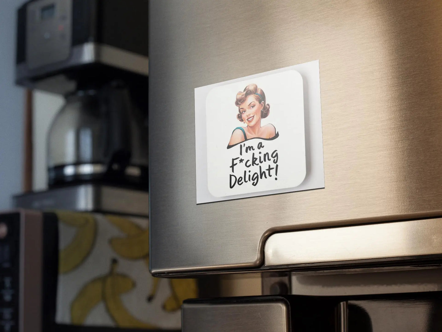 An "I'm a fucking delight" snarky magnet from Mirabilia Boutique, featuring an illustration of a smiling woman and the text "I'm a f*ing delight!", is attached to the door of a stainless steel fridge. In the background, there is a coffee maker and banana-themed wallpaper, adding to the humorous vibe.